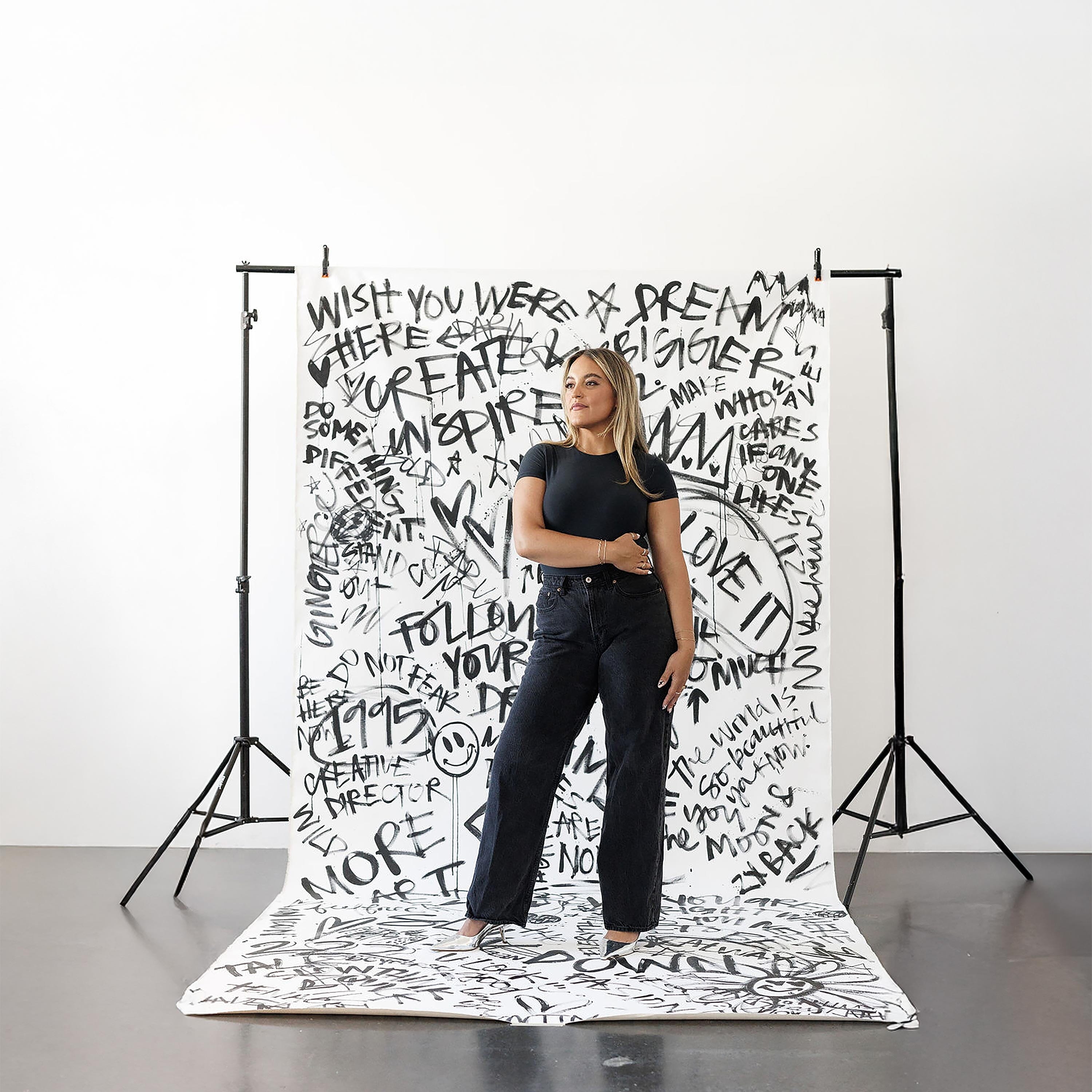 Artist Ginger Hamilton standing confidently in front of a bold, black-and-white graffiti-style backdrop featuring hand-lettered inspirational phrases.